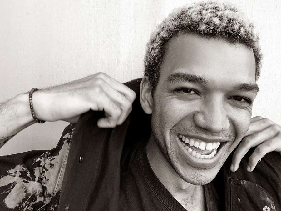 A cheerful man with curly hair smiling while adjusting his jacket. — Justice Smith Wallpaper