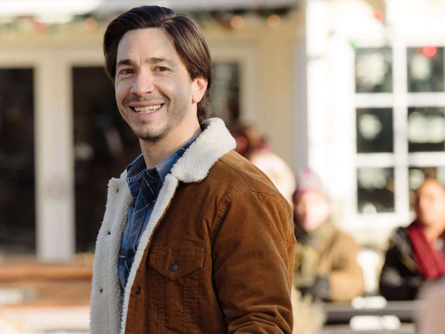 En mann i en brun shearling-jakke smiler varmt utendørs nær en festlig bygning. — Justin Long Bakgrunner