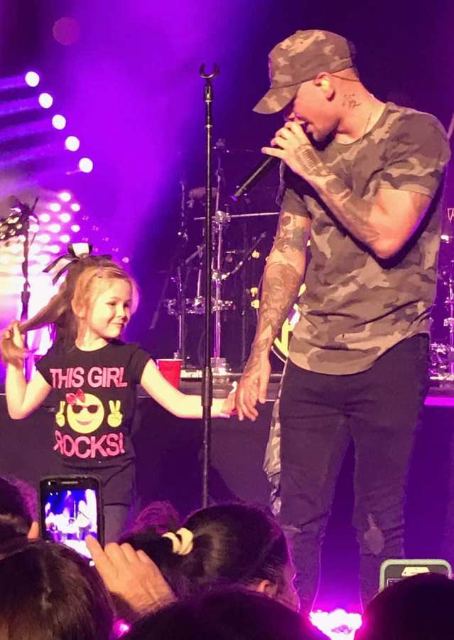 Ein Sänger und ein junges Mädchen halten sich auf einer Konzertbühne unter lila Lichtern an den Händen. — Kane Brown Bilder