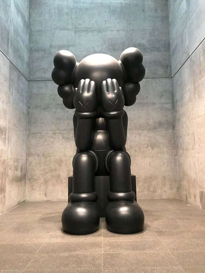 Large black KAWS sculpture covering its eyes in a concrete room. — Kaws Artwork Wallpaper