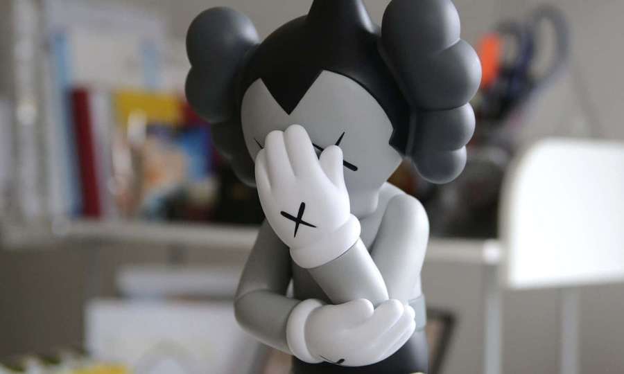 Gray and white KAWS figurine covering its face indoors. — Kaws Astro Boy Wallpaper