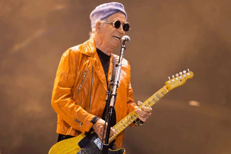 A musician in an orange jacket and purple beanie plays guitar and sings on stage. — Keith Richards Wallpaper