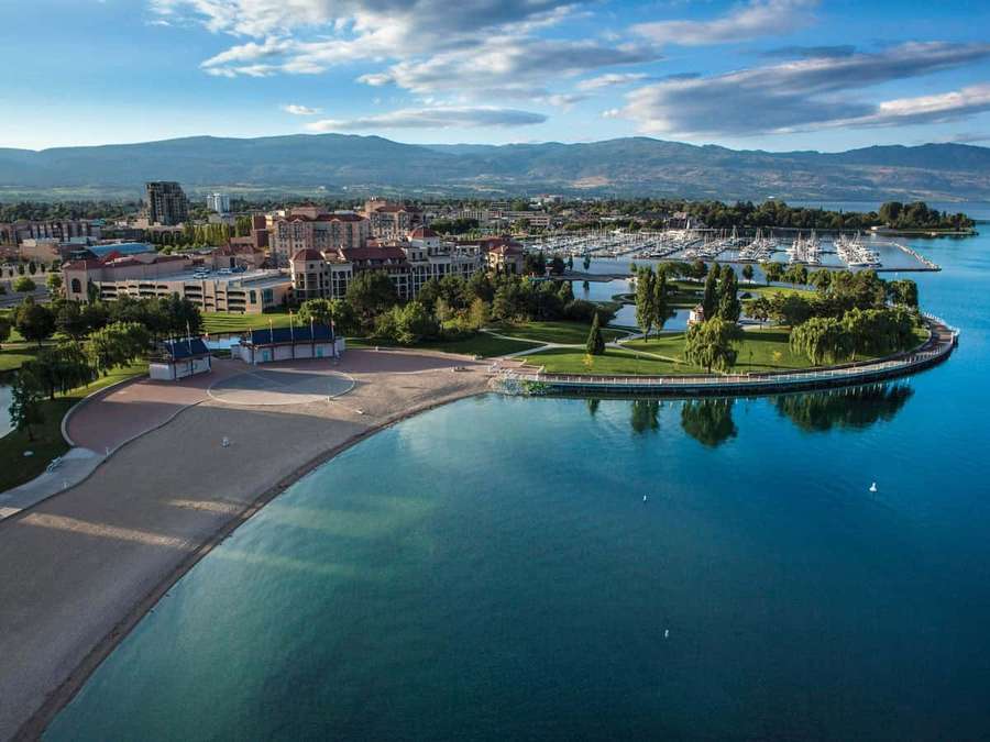 Coastal town with marina boats, green parks, and mountains under a partly cloudy sky. — Kelowna Wallpaper