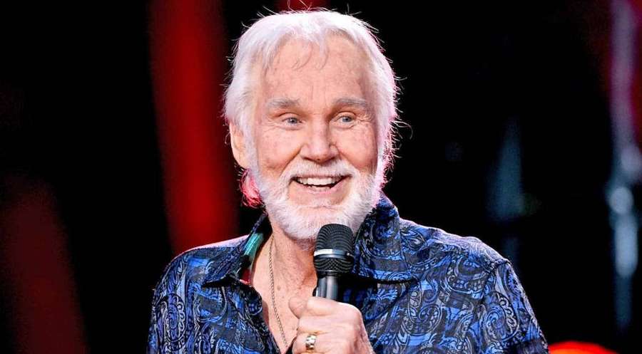 Ein älterer Mann mit weißen Haaren und Bart lächelt, während er ein Mikrofon auf der Bühne hält. — Kenny Rogers Bilder
