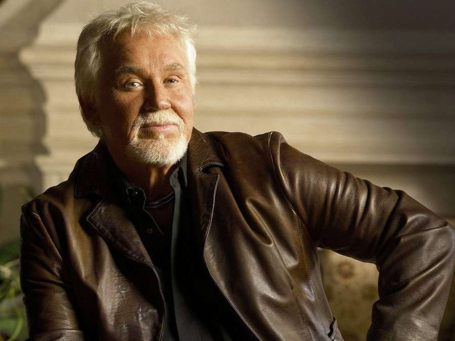 Un uomo anziano con capelli e barba bianchi in una giacca di pelle marrone sorride calorosamente. — Kenny Rogers Sfondi