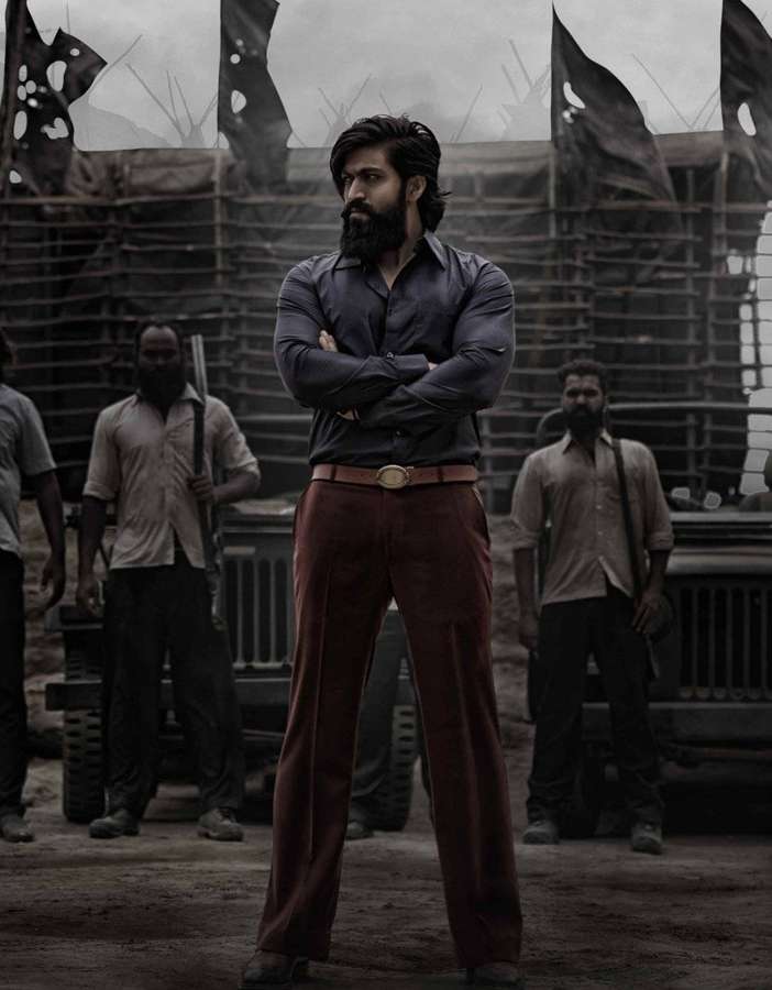 A bearded man in a dark shirt and red pants stands arms crossed in a war-torn urban setting. — Kgf Chapter 2 Wallpaper