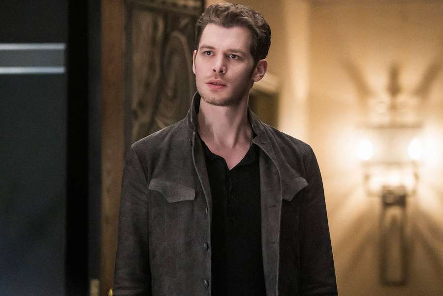 Ein Mann in einer dunklen Jacke steht in einem warm beleuchteten Flur mit verzierten Wandleuchtern. — Klaus Mikaelson Wallpaper
