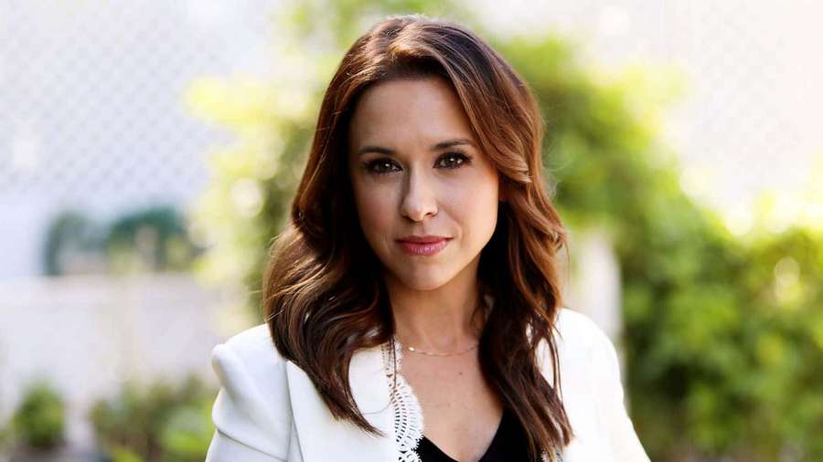 Woman in white blazer and black top outdoors with greenery behind her. — Lacey chabert Wallpaper