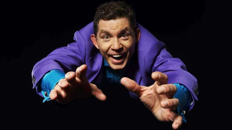 Hombre con chaqueta morada extendiendo la mano hacia adelante con expresión emocionada sobre fondo negro — Lee Evans Fondos de pantalla