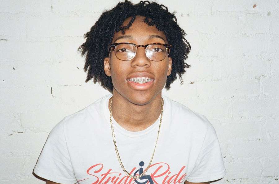 Young person with curly hair glasses and braces wearing a white t-shirt and gold chain. — Lil Tecca Background