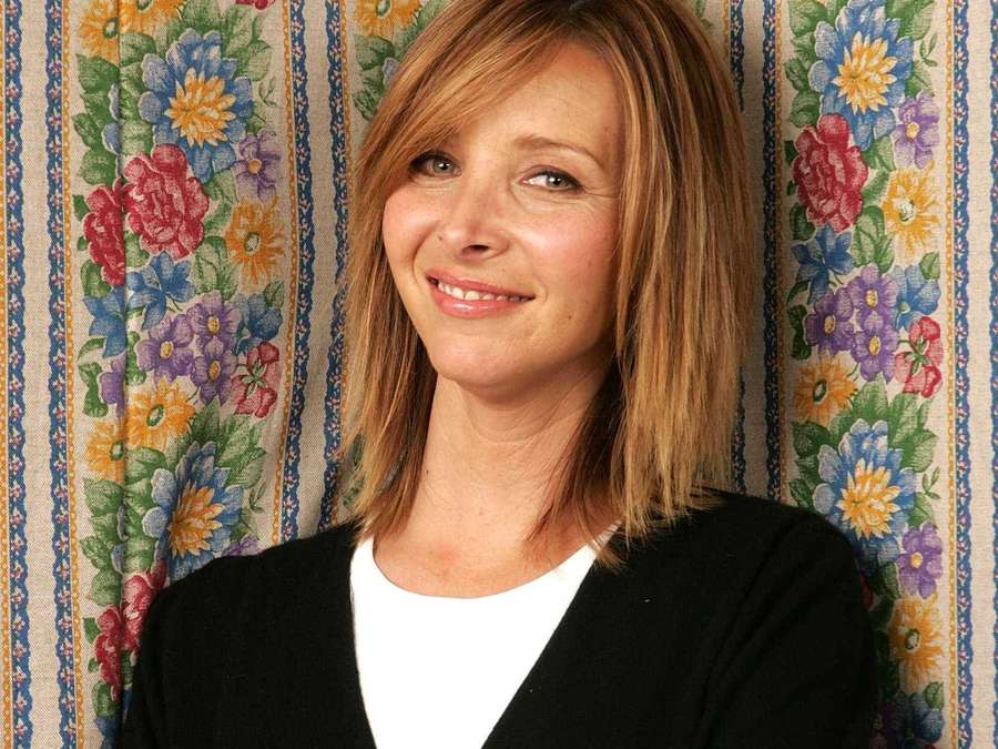Eine lächelnde Frau in einem schwarzen Cardigan und weißem Hemd vor einer Blumentapete. — Lisa kudrow Bilder