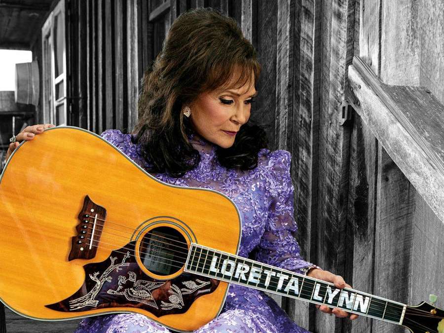 Loretta Lynn holding a guitar in front of wooden siding. — Loretta Lynn Wallpaper