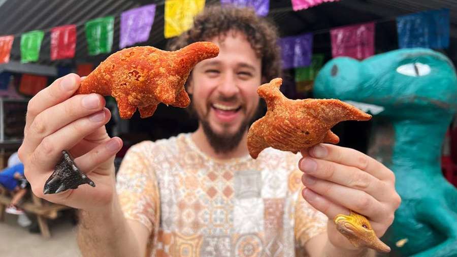 Man holding dinosaur-shaped fried snacks at a colorful outdoor festival. — Luisito Comunica Wallpaper