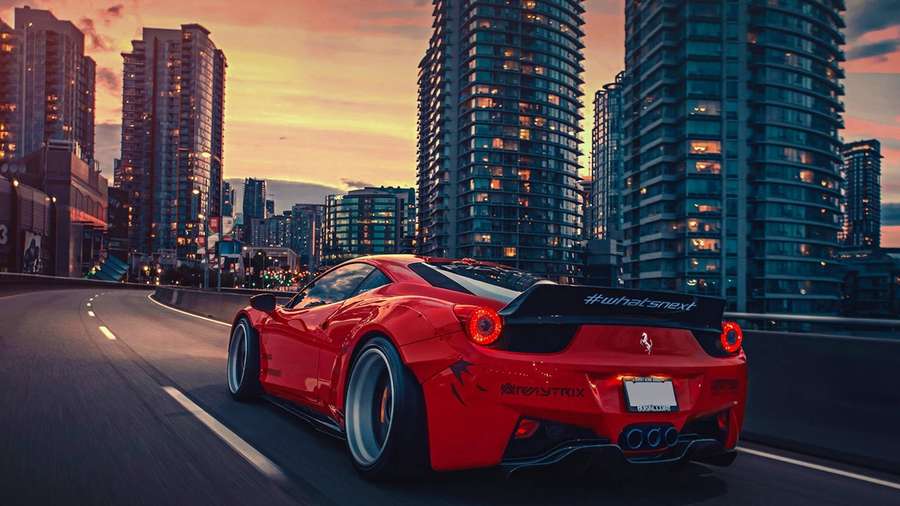 Ferrari rossa che sfreccia in città al tramonto con grattacieli illuminati sullo sfondo — Lusso Sfondi