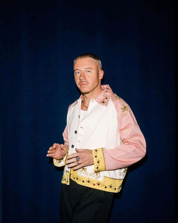 Man wearing pink and white embroidered jacket against blue background — Macklemore Wallpaper