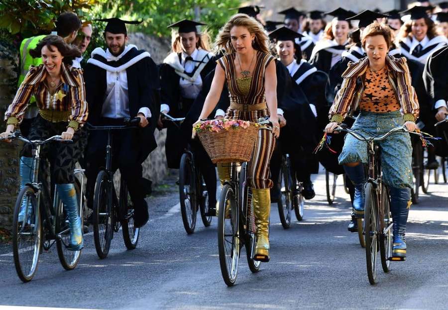 Students in graduation gowns ride bicycles down a sunny street. — Mamma Mia Background