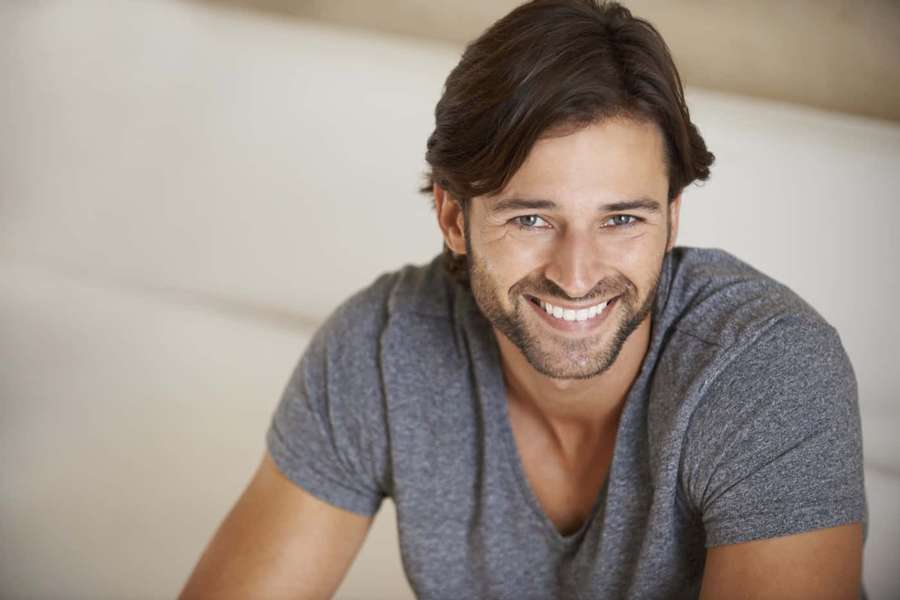 A man with a beard smiles while leaning on a couch indoors. — Man Picture