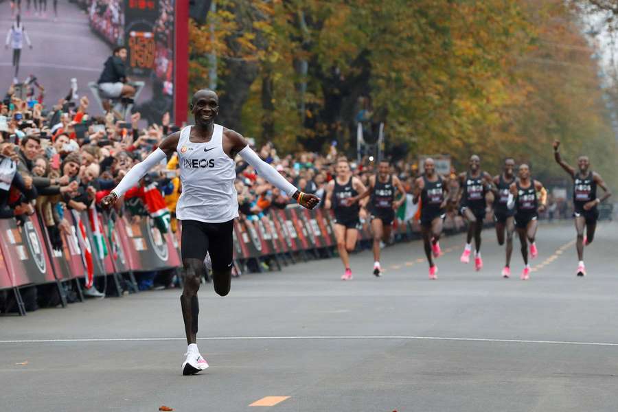 A smiling runner crosses a street finish line with cheering crowds and autumn trees in the background. — Marathon Background