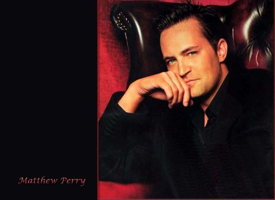 Matthew Perry dalam setelan hitam duduk di kursi kulit merah dengan ekspresi penuh pemikiran. — Matthew perry Wallpaper