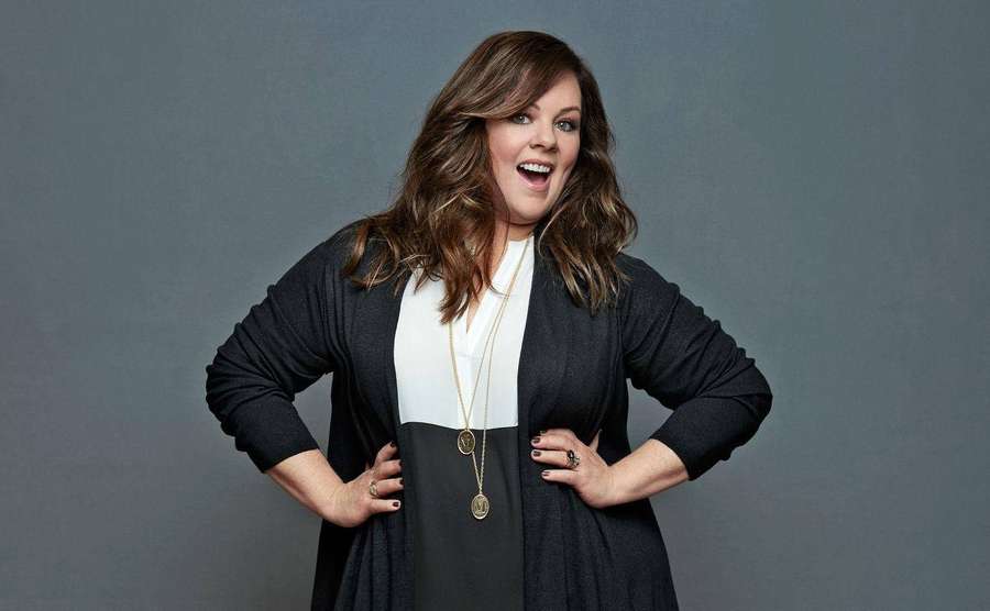 A smiling woman in a black cardigan and white top poses with hands on hips. — Melissa mccarthy Background