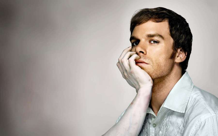 A man in a light shirt rests his chin on his hand looking thoughtfully at the camera. — Michael c. hall Wallpaper