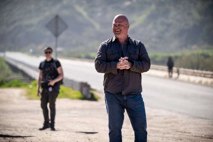 Ein kahlköpfiger Mann lacht, während er am Straßenrand steht und eine andere Person hinter ihm geht. — Michael Chiklis Bilder