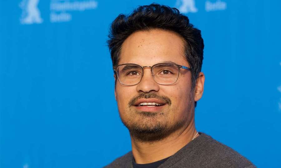 Man with glasses and beard smiling at camera against blue event backdrop. — Michael Peña Wallpaper