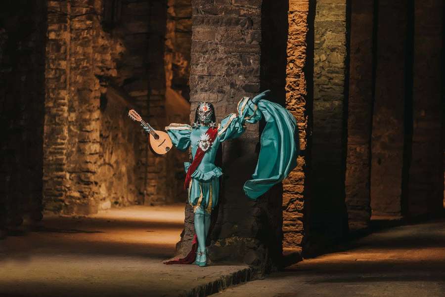 A masked performer in teal costume plays a lute in ancient stone ruins. — Middle Ages Wallpaper