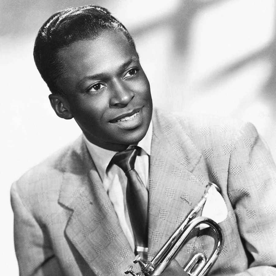 En smilende svart jazztrompetist i en mønstret dress som holder instrumentet sitt. — Miles Davis Bakgrunner