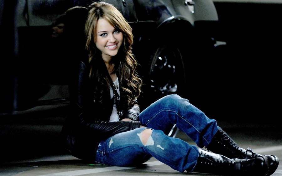 Young woman in ripped jeans and boots smiling beside a car at night. — Miley Wallpaper