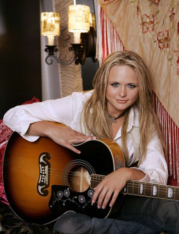 Blonde woman holding acoustic guitar in warmly lit room with floral tapestry and candle sconces. — Miranda Lambert Wallpaper