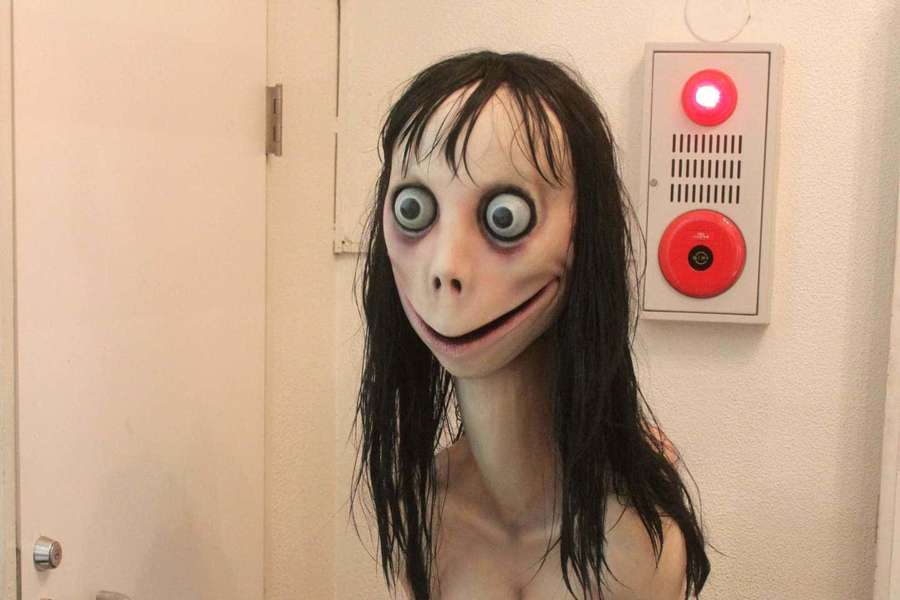 Griezelig animatronisch masker met grote ogen en sinistere grijns bij een rood verlichte deurintercom. — Momo Achtergronden