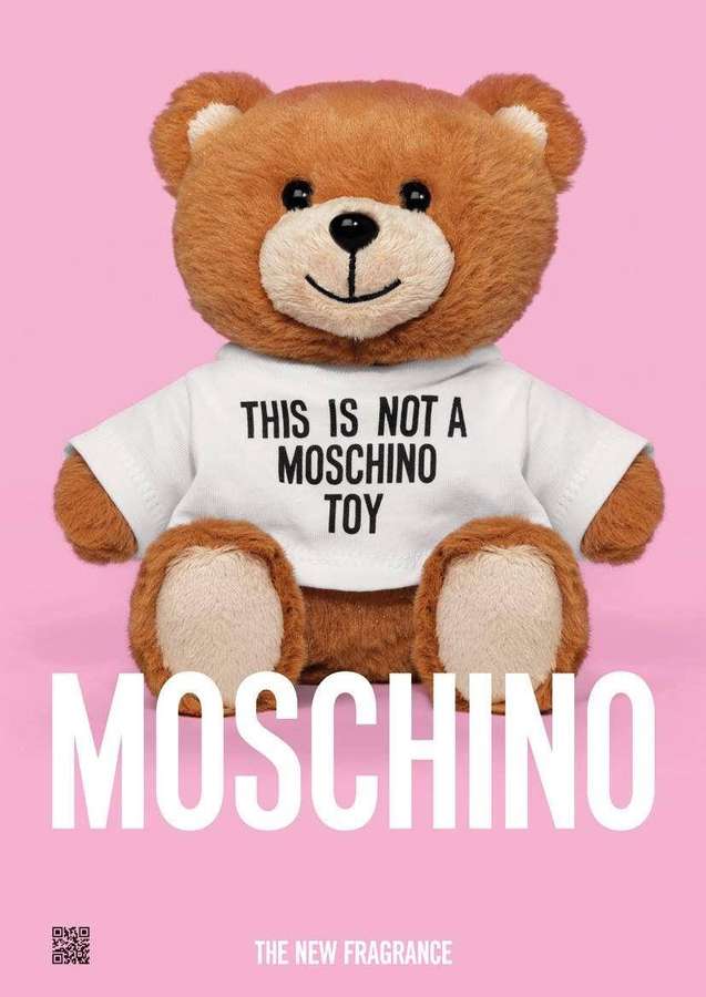 A brown teddy bear wearing a t-shirt that reads 'This is not a Moschino toy' against a pink background. — Moschino Wallpaper