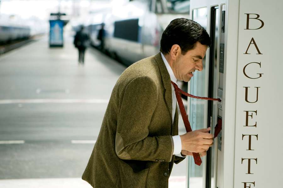 Man in tweed jacket and red tie trying to open a baguette vending machine at a train station. — Mr Bean 4K Wallpaper