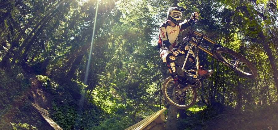 Mountain biker airborne over wooden ramp in sun-dappled forest. — Mtb Background
