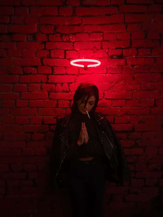 Persona con chaqueta negra fumando cigarrillo bajo halo de neón rojo contra pared de ladrillos. — Mujer rezando Fondos de pantalla