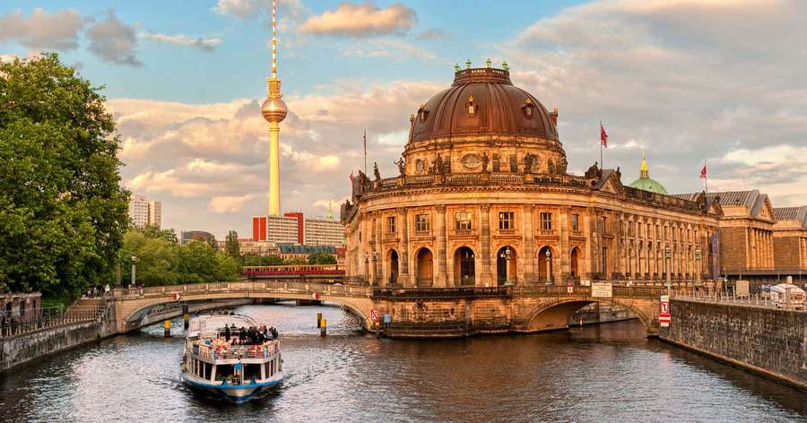Tour boat glides under a bridge beside Berlin's Reichstag and TV tower at sunset. — Museum Island Wallpaper
