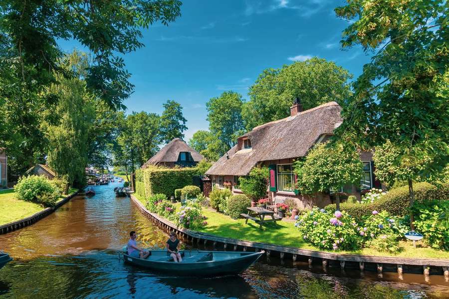 Two people rowing a boat through a picturesque canal village with thatched houses and blooming flowers. — Netherlands Picture