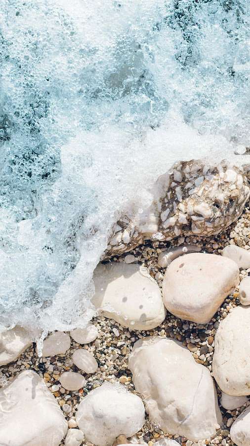 Meereswellen, die über glatte Kieselsteine an einem sonnigen Strand brechen. — Nettes Strand iPhone Wallpaper
