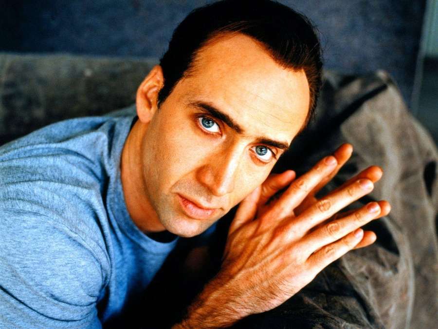 A man with intense blue eyes rests his hand on his cheek in a close-up portrait. — Nicolas Cage Background