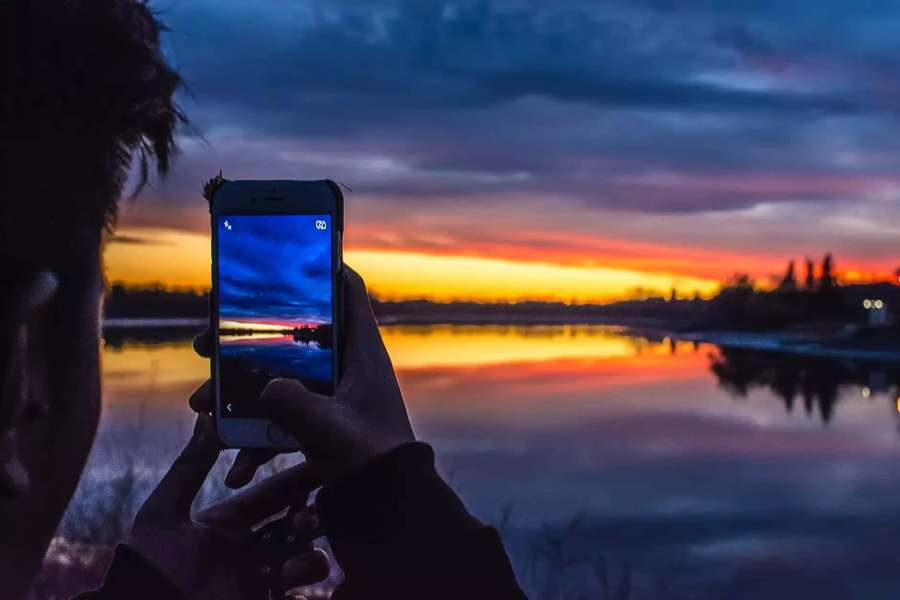 Person holding phone to photograph sunset over lake at dusk — Night Mode Wallpaper