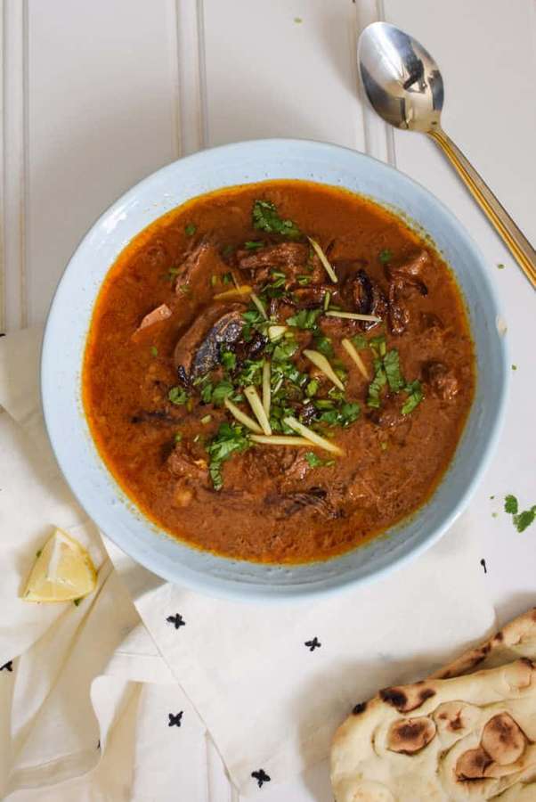 A bowl of lamb curry with naan bread and lemon wedge on a wooden table. — Nihari Picture