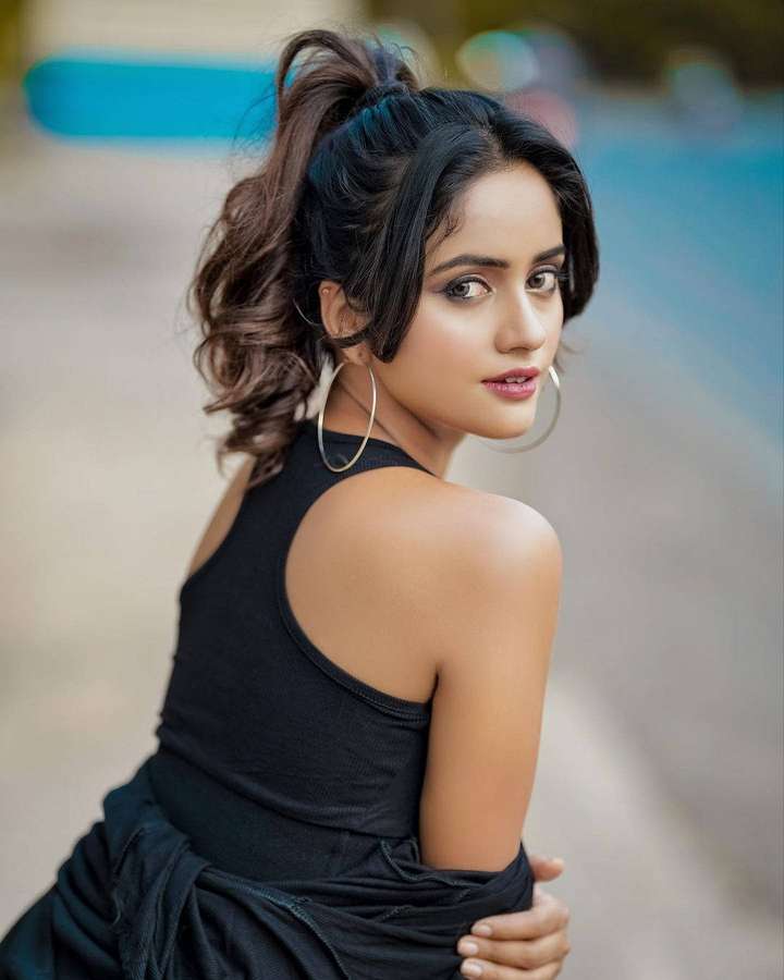 A woman in a black top and hoop earrings turns her head over her shoulder outdoors. — Nisha Wallpaper