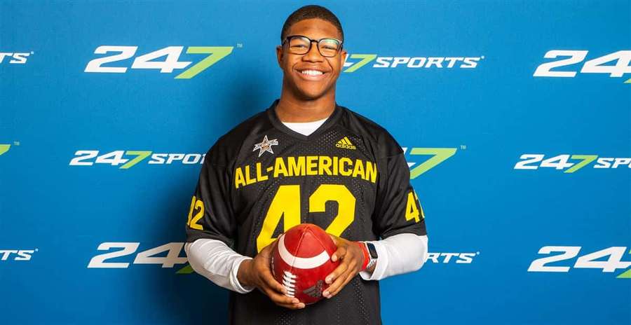 Young football player in All-American jersey holding a football in front of blue branded wall. — Nolan Smith Wallpaper