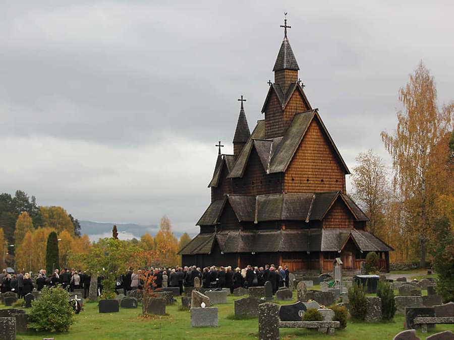 Una iglesia de madera rodeada de lápidas con personas reunidas para una ceremonia. — Noruega Fondos de pantalla
