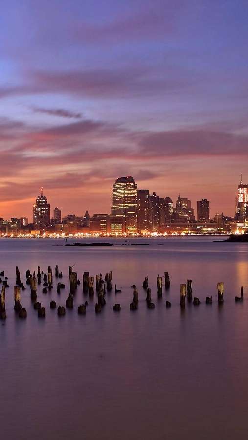 Horizonte de la ciudad al atardecer con aguas tranquilas y pilotes de madera en primer plano. — Nueva Jersey Fondos de pantalla