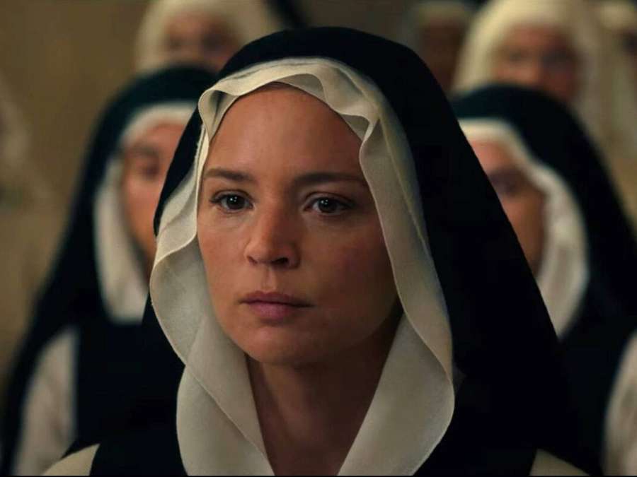 A nun in black and white habit looks solemnly forward in a dimly lit room with blurred nuns behind her. — Nun Wallpaper