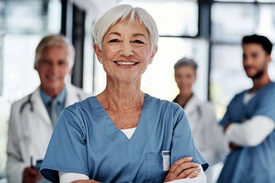 Smiling senior nurse in blue scrubs with medical team in background. — Nursing Picture