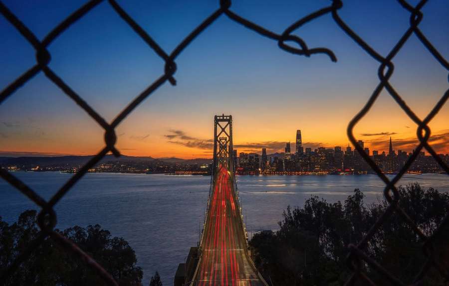 Golden Gate Bridge at sunset viewed through a chain-link fence with city lights and traffic streaks. — Oakland Wallpaper