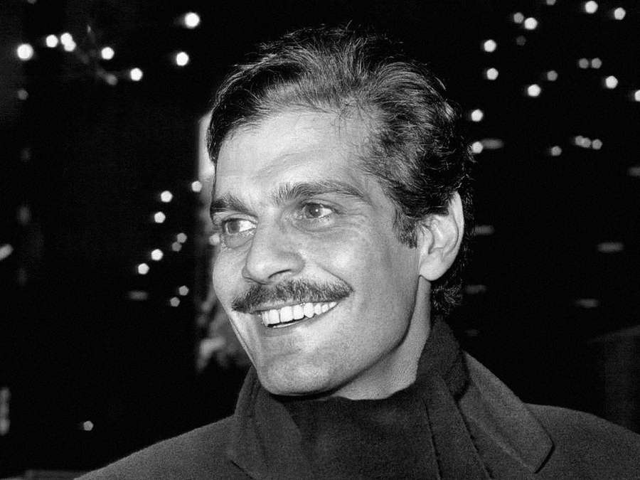 Ein lächelnder Mann mit Schnurrbart auf einem Schwarz-Weiß-Foto bei Nacht mit Stadtlichtern hinter ihm. — Omar Sharif Bilder